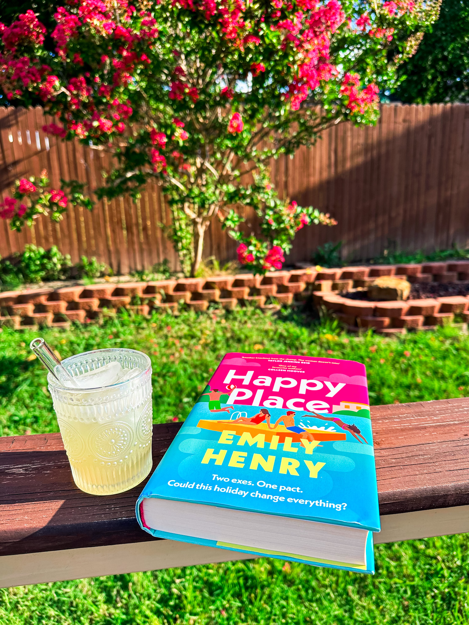 A hardcover copy of the book 'Happy Place' by Emily Henry sits on a wooden railing beside a glass of lemonade with a glass straw. In the background, a green lawn, a wooden fence, and a blooming crepe myrtle tree with pink flowers are visible, creating a serene and summery outdoor setting.