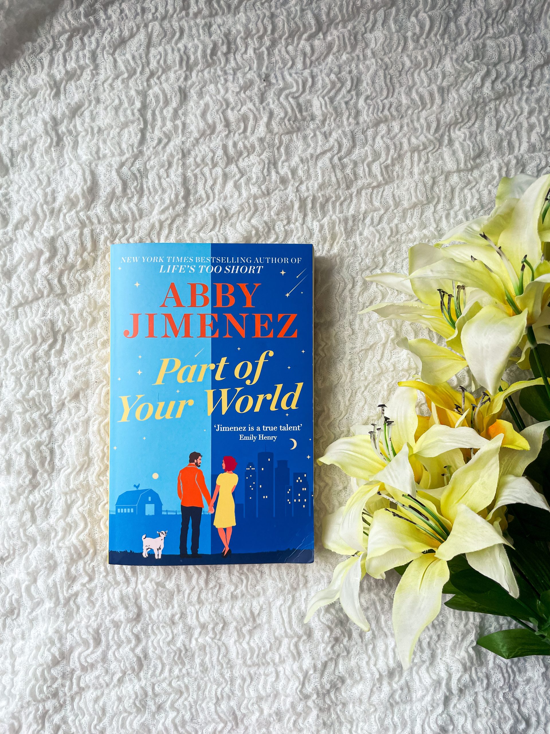 A copy of 'Part of Your World' by Abby Jimenez resting on a textured white blanket, with a bouquet of light yellow lilies to the right of the book.