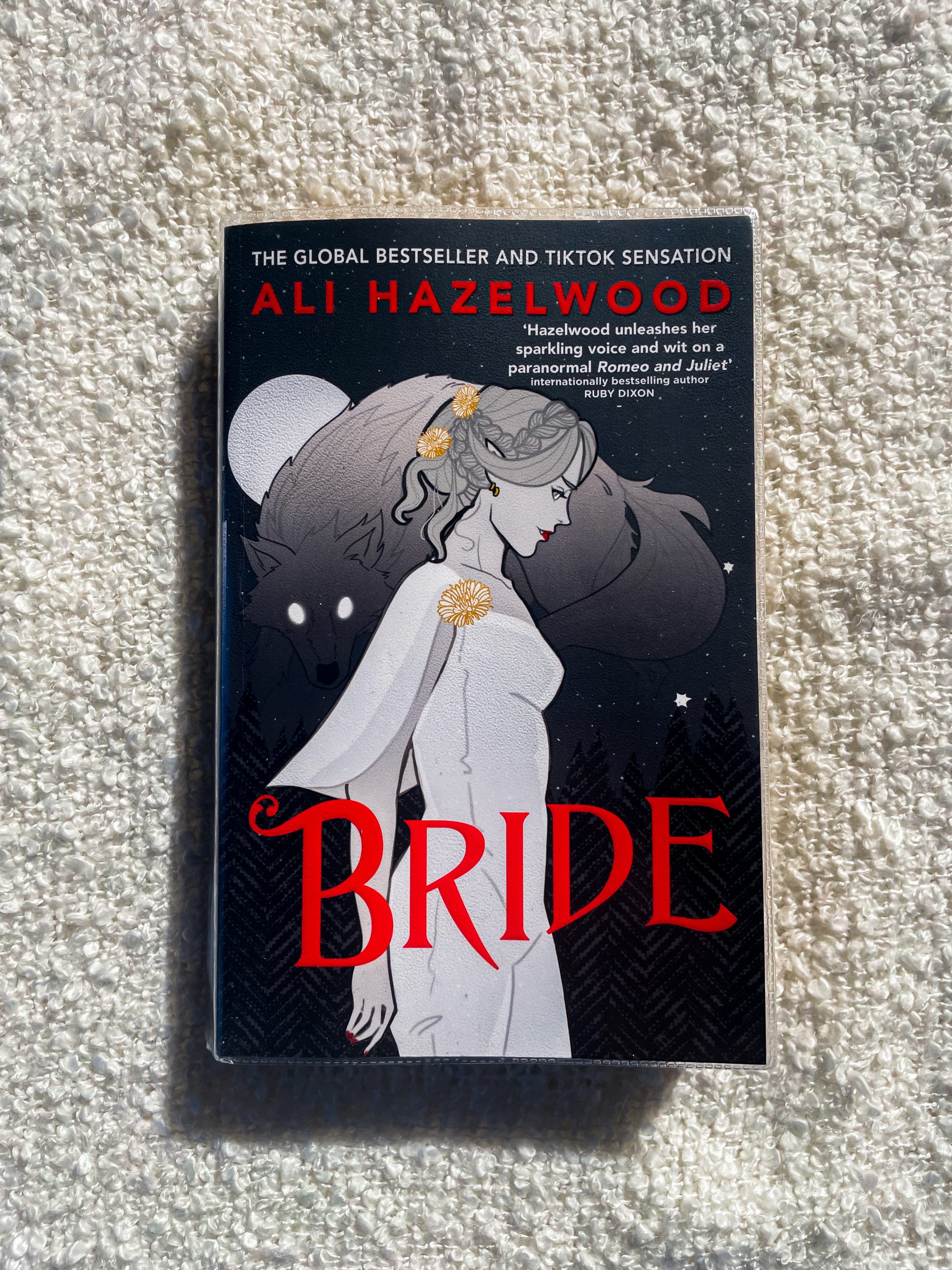 Cover of the book Bride by Ali Hazelwood. The cover features an illustration of a woman in a white dress, adorned with golden flower accessories, standing against a dark, starry background. Behind her is a shadowy figure of a wolf. The title BRIDE is in bold red letters, and the author's name ALI HAZELWOOD is displayed prominently at the top.