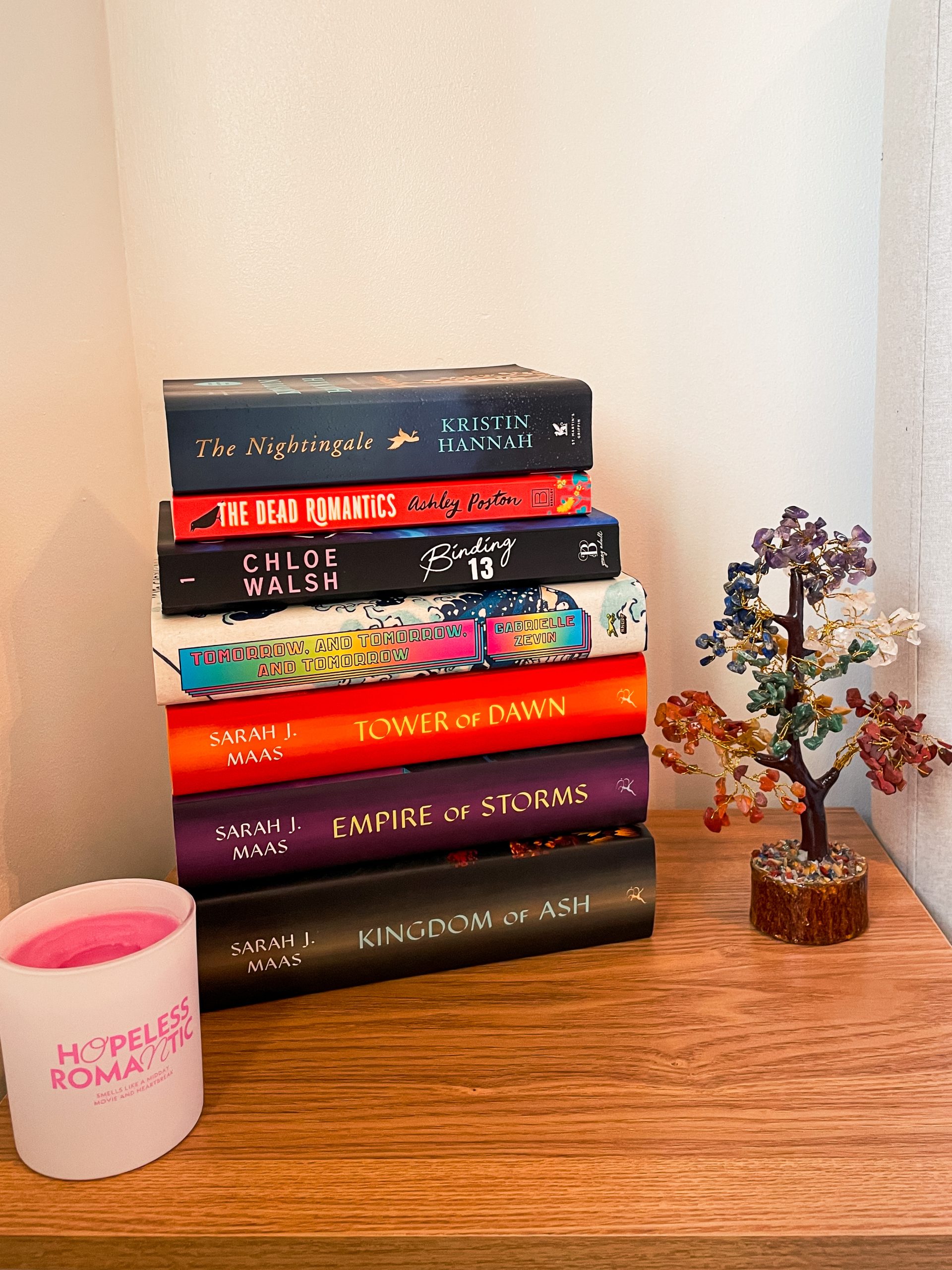 Stack of popular books on a wooden surface featuring 'The Nightingale,' 'The Dead Romantics,' 'Binding 13,' 'Tomorrow, and Tomorrow, and Tomorrow,' and Sarah J. Maas's Throne of Glass series, with a pink 'Hopeless Romantic' candle and a decorative gemstone tree.