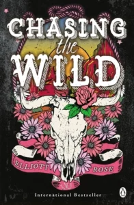 Book cover of Chasing the Wild by Elliott Rose featuring a decorated bull skull surrounded by pink flowers and roses, with a desert landscape and sunset in the background.