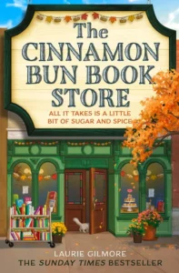 Book cover of The Cinnamon Bun Bookstore by Laurie Gilmore featuring a cozy green storefront decorated with pumpkins, autumn leaves, and a cat at the door, a perfect pick for readers searching for autumn romance books.